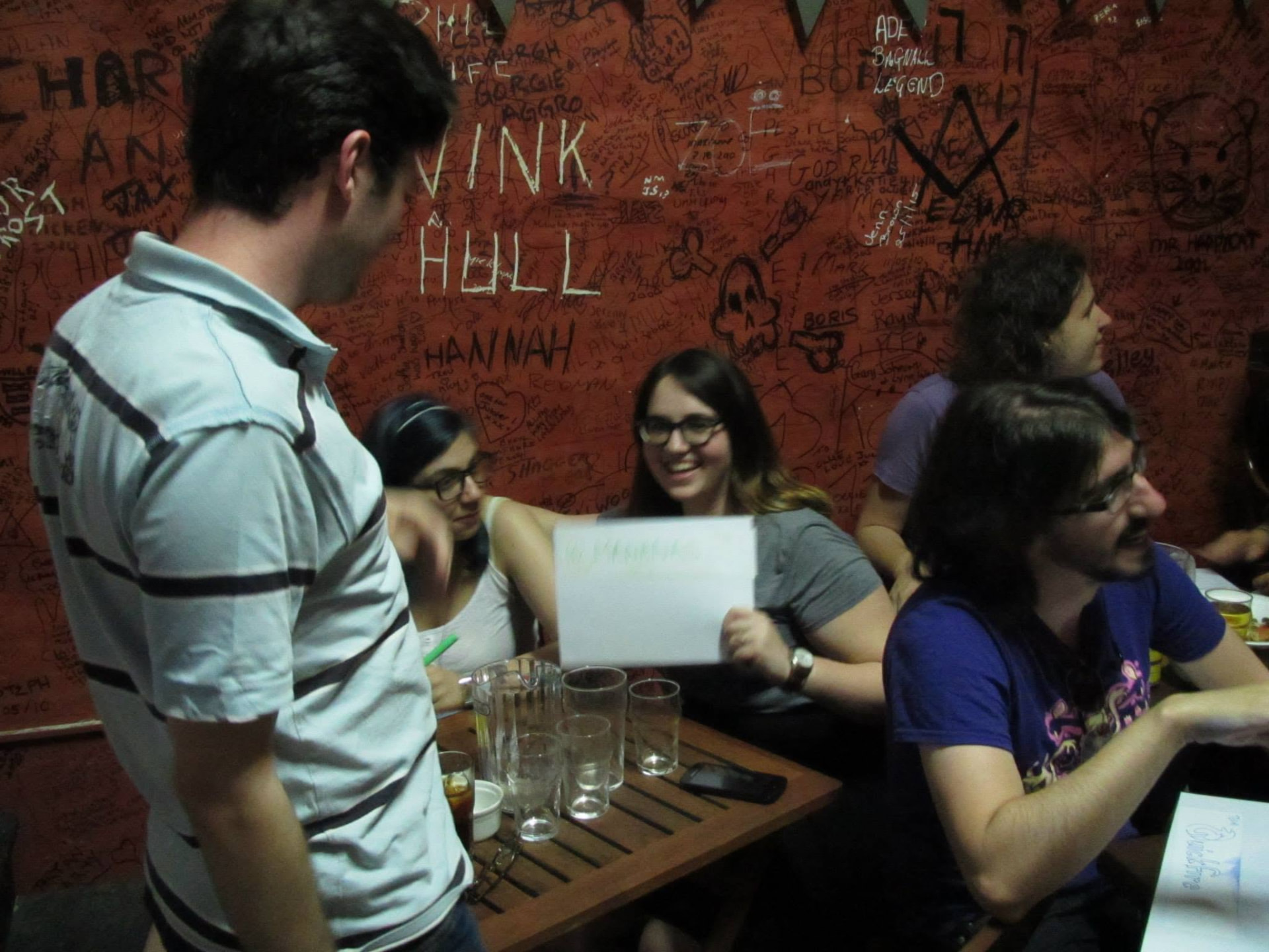 People seated at a table in front of a wall covered in handwritten signatures, one holding up a sheet of paper